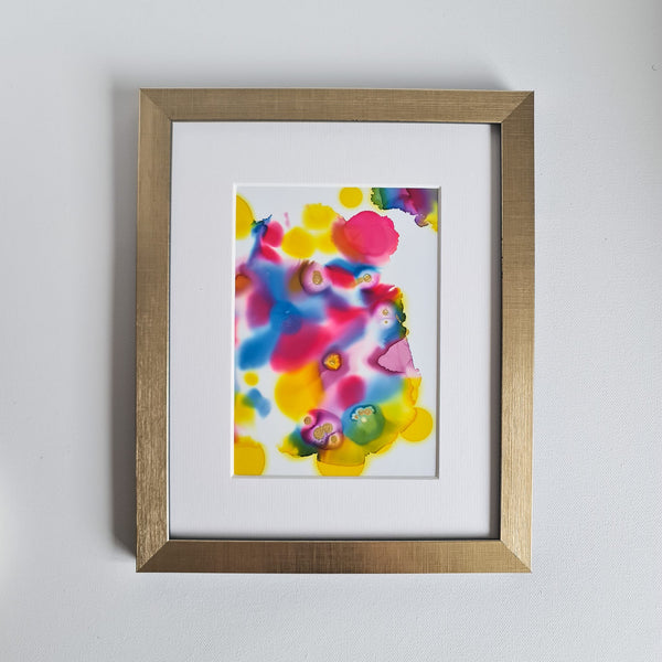 "Warhol Two, 2018" is an abstract alcohol ink painting featuring bold, vibrant colors of red, yellow, and blue. Swirls and splashes create a sense of movement. Mounted on a white background, and sitting in a gold frame.
