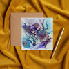 Square greeting card with abstract purple and blue floral design beside a kraft envelope and gold pen on golden velvet fabric