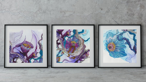 Three framed abstract paintings with floral and organic shapes in purple, blue, and yellow hues on a concrete floor.
