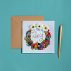 Square greeting card with colorful bead and floral wreath design encircling the word "Joy," paired with a kraft envelope and gold pen on turquoise fabric