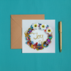 Square greeting card with colorful bead and floral wreath surrounding the word Joy, paired with kraft envelope and gold pen on teal fabric