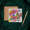 Square greeting card with abstract pink and blue floral artwork, paired with a brown envelope and gold pen on dark green fabric