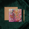 Square greeting card with colorful abstract floral embroidery and bead details beside a kraft envelope and gold pen on dark green fabric