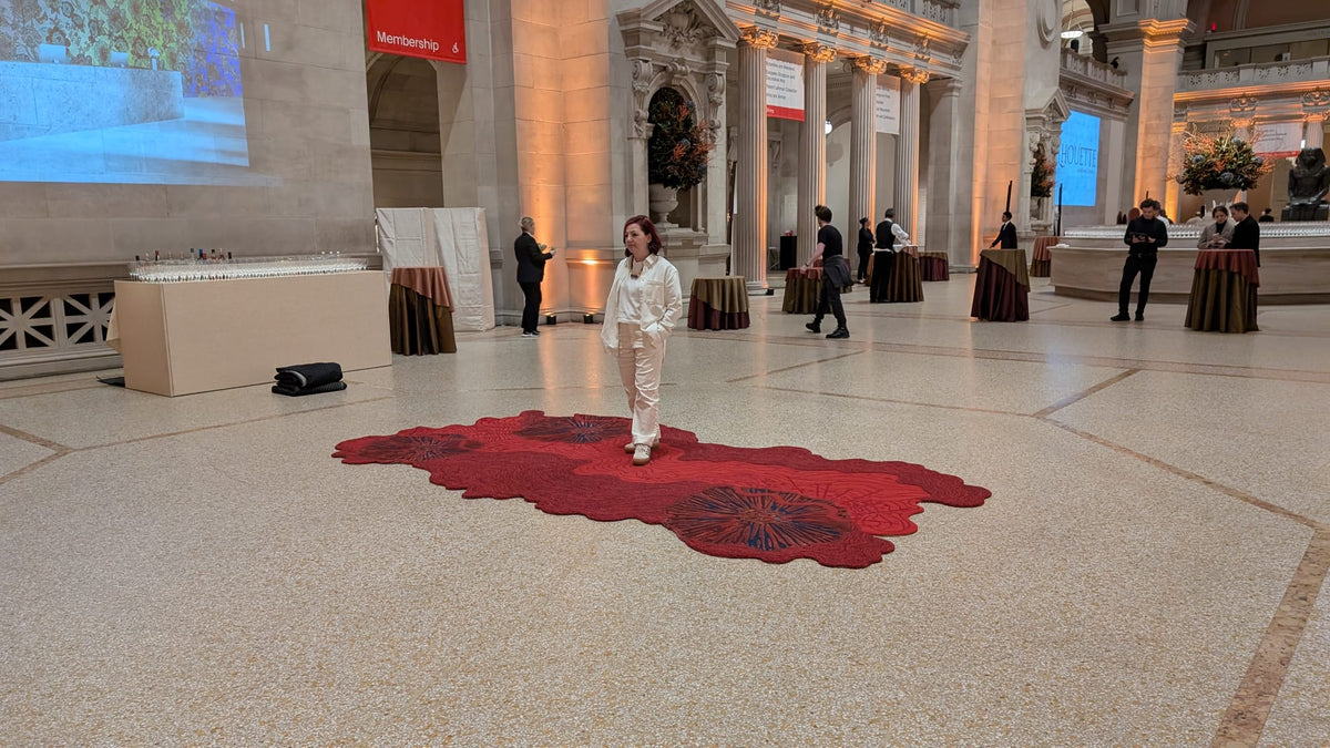 Person walking on a large red textile artwork displayed on the floor of a grand museum hall with columns and sculptures