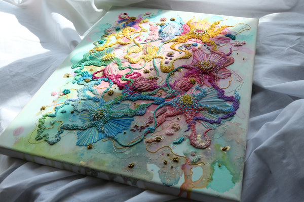 Embroidery artwork with pastel-colored floral designs and beadwork displayed on a canvas resting on white fabric