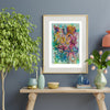 Viola Five 2022 abstract floral art print in gold frame on blue wall above wooden table with plants.
