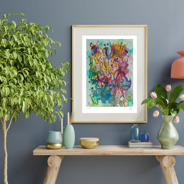 Viola Five 2022 abstract floral art print in gold frame on blue wall above wooden table with plants.