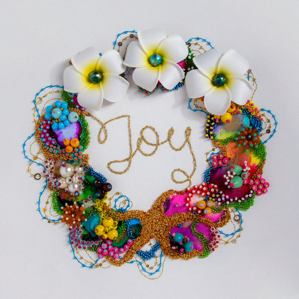Colorful beaded and embroidered circular artwork with three white flowers surrounding the word "joy" on a white background