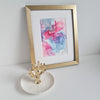 Abstract alcohol ink artwork "Aphrodite Plays, 2018" in gold frame with pink, purple, and blue hues on white surface