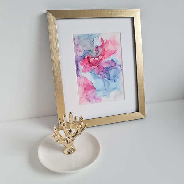 Abstract alcohol ink artwork "Aphrodite Plays, 2018" in gold frame with pink, purple, and blue hues on white surface