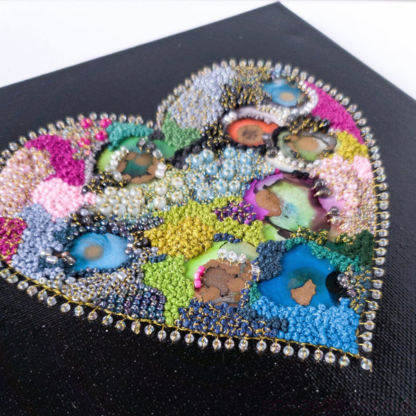Close-up of a colorful embroidered heart artwork with beads and alcohol ink on black canvas.