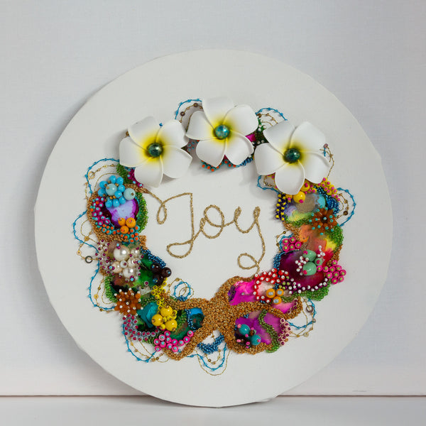 Round coral wreath artwork with colorful beads, embroidery, faux flowers, and embroidered word "Joy" in gold thread.