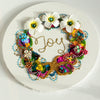 Round coral-inspired wreath with colorful beads, embroidery, faux flowers, and "Joy" embroidered in gold thread.