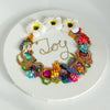 Coral wreath artwork with beads, embroidery, faux flowers, and gold thread spelling Joy on round white canvas.