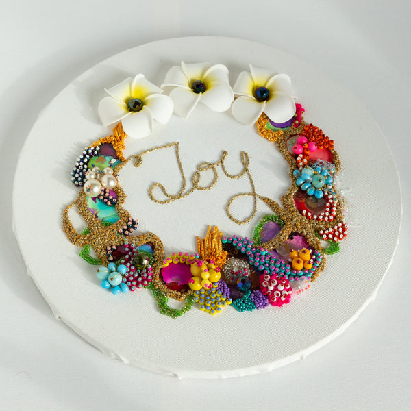 Coral wreath artwork with beads, embroidery, faux flowers, and gold thread spelling Joy on round white canvas.