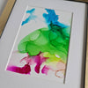 Abstract alcohol ink artwork with vibrant green, blue, and pink hues on Yupo paper in white mount