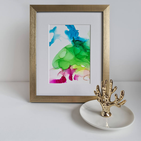 Abstract alcohol ink painting Dionysus 2018 in gold frame with white mount displayed on white surface next to gold coral ring holder