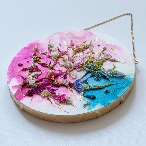 Round mixed media artwork with alcohol ink, hand embroidery, seed beads, and gold leafed edges hanging by a gold chain
