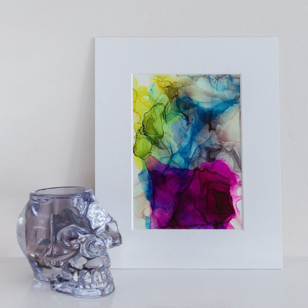 Abstract alcohol ink painting "Hera, 2018" with vibrant colors, mounted in white frame, displayed beside a glass skull decor.