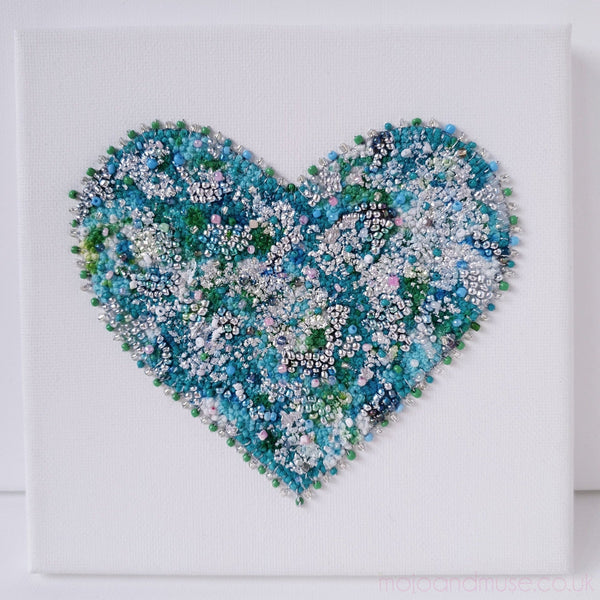 Close-up of a textured mint green and blue beaded heart embroidery on white canvas, handmade art piece.