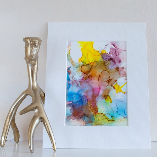 Abstract alcohol ink painting titled Odin's Glory 2018 with vibrant yellow, pink, blue, and brown hues on white mount.