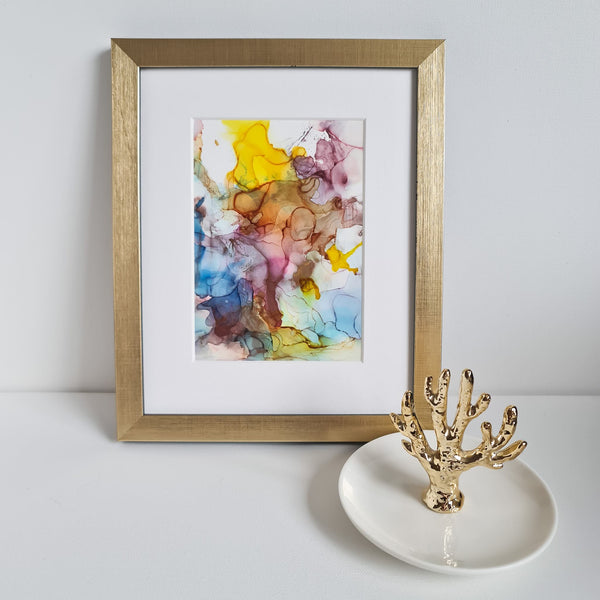 Abstract alcohol ink painting with vibrant colors, mounted in white mat within a gold frame on a white surface.