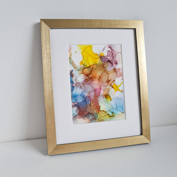 Abstract alcohol ink painting titled Odin's Glory with vibrant colors, mounted in white with a gold frame on a white surface.