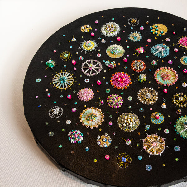Round mixed-media artwork depicting colorful diatoms with beads and sequins on black canvas, titled Sea Jewels in the Snow Two.