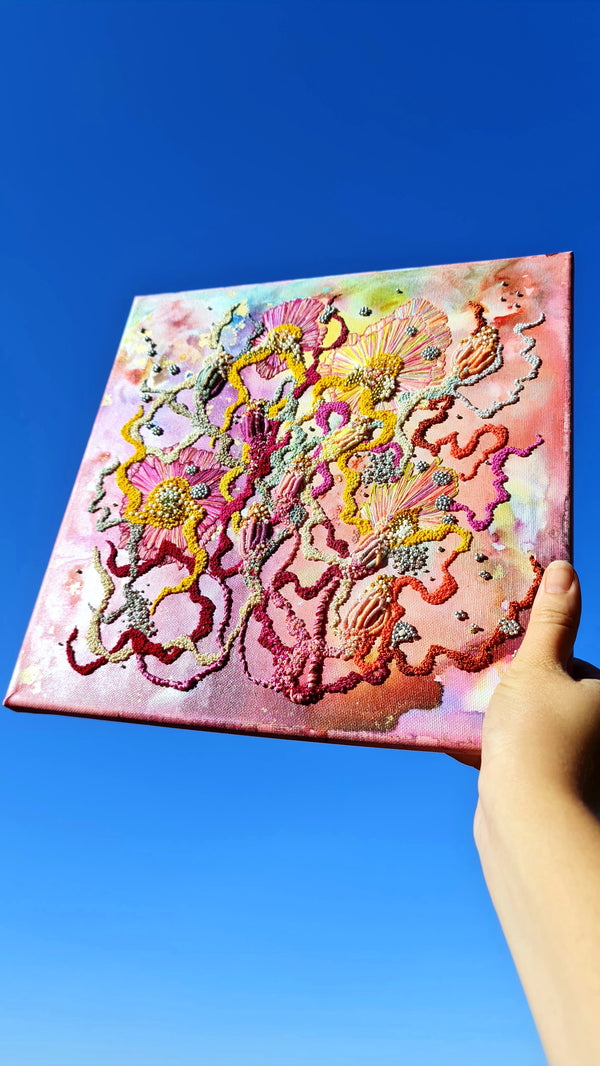 Hand-embroidered abstract art with alcohol ink, seed beads, and metallic threads on canvas held against clear blue sky