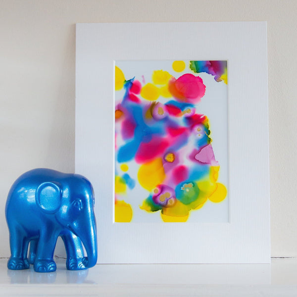 Abstract colorful alcohol ink artwork titled Warhol Two 2018 in white mount, displayed beside a small blue elephant figure.